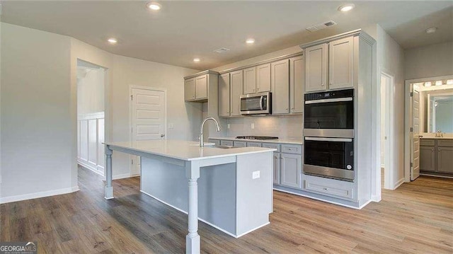 kitchen featuring decorative backsplash, light hardwood / wood-style flooring, an island with sink, gray cabinetry, and stainless steel appliances