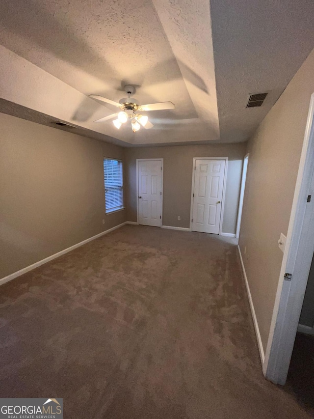 unfurnished bedroom featuring ceiling fan, dark carpet, a raised ceiling, and a textured ceiling