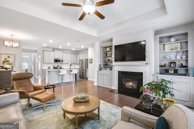 living room with a raised ceiling, built in shelves, ceiling fan with notable chandelier, and hardwood / wood-style flooring