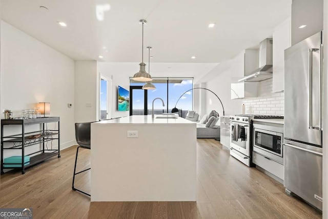 kitchen featuring hanging light fixtures, wall chimney range hood, a center island with sink, decorative backsplash, and stainless steel appliances