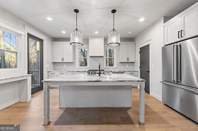 kitchen featuring a center island with sink, custom exhaust hood, high end fridge, light stone countertops, and white cabinets