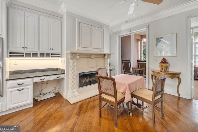 dining area featuring built in desk, light hardwood / wood-style floors, ceiling fan, crown molding, and a tiled fireplace
