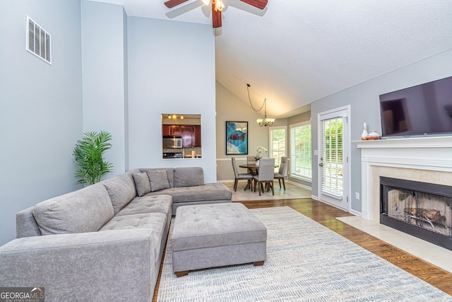 living room featuring ceiling fan with notable chandelier, a textured ceiling, wood-type flooring, high vaulted ceiling, and a tiled fireplace