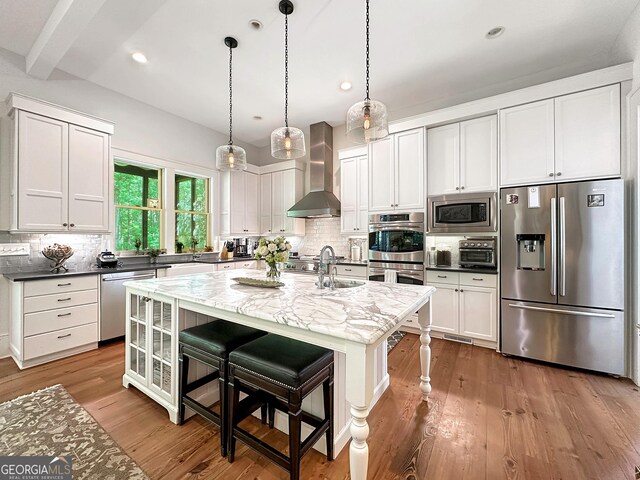 kitchen featuring wall chimney range hood, dark stone countertops, a center island with sink, a breakfast bar, and appliances with stainless steel finishes