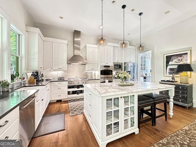 kitchen featuring appliances with stainless steel finishes, wall chimney range hood, pendant lighting, white cabinets, and a center island