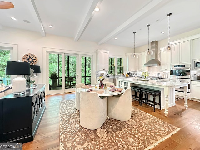 kitchen with a breakfast bar area, a kitchen island with sink, wall chimney range hood, white cabinets, and beamed ceiling