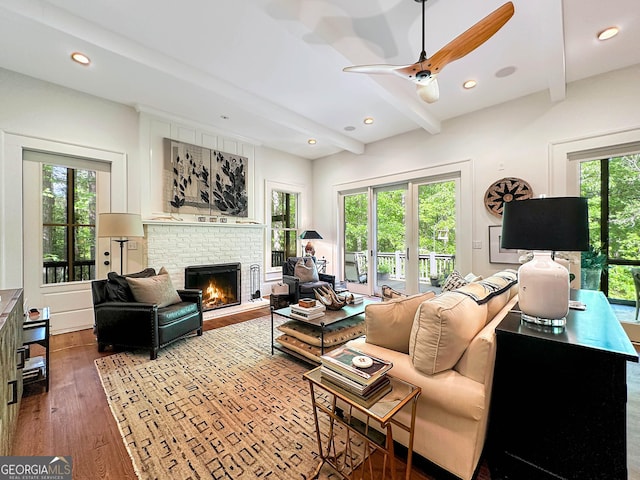 living room featuring a brick fireplace, beam ceiling, wood-type flooring, and ceiling fan