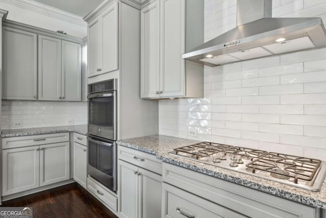 kitchen with tasteful backsplash, wall chimney range hood, stainless steel appliances, ornamental molding, and light stone counters