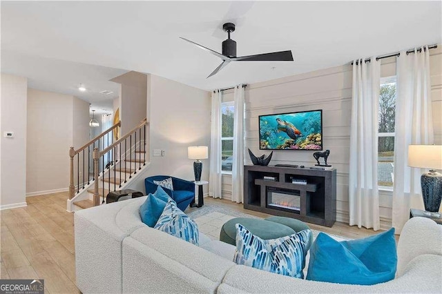 living area featuring wood finished floors, a ceiling fan, baseboards, stairs, and a glass covered fireplace