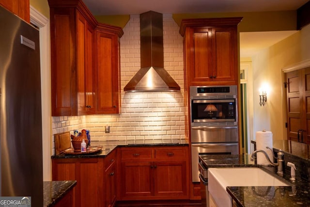 kitchen featuring oven, dark stone counters, fridge, and wall chimney range hood