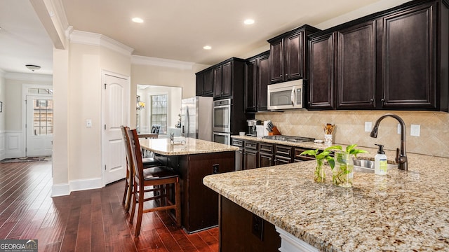 kitchen with a breakfast bar, a center island with sink, stainless steel appliances, ornamental molding, and light stone countertops