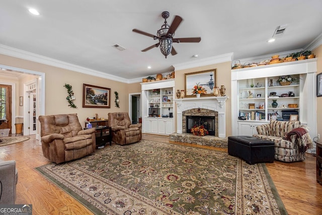 living room with ceiling fan, wood-type flooring, crown molding, a stone fireplace, and built in shelves