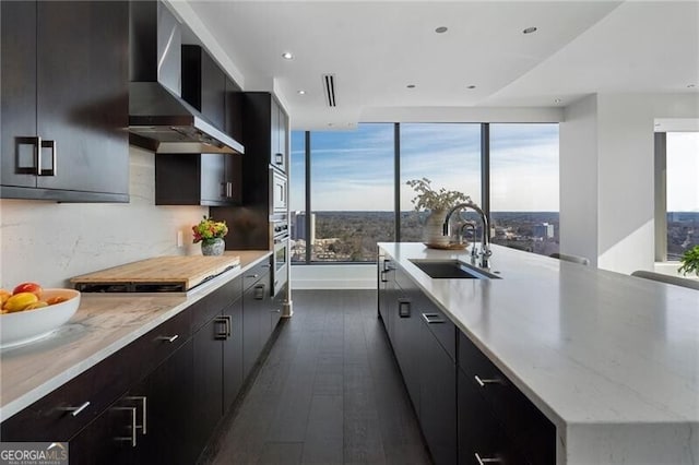 kitchen with wall chimney range hood, a center island with sink, cooktop, sink, and dark hardwood / wood-style flooring