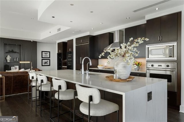 kitchen featuring wall chimney range hood, stainless steel appliances, an island with sink, sink, and a breakfast bar