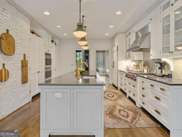 kitchen with wall chimney range hood, white cabinets, hanging light fixtures, and an island with sink