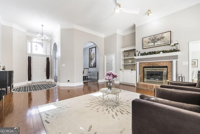 living room with ceiling fan with notable chandelier, dark hardwood / wood-style flooring, built in features, a brick fireplace, and crown molding