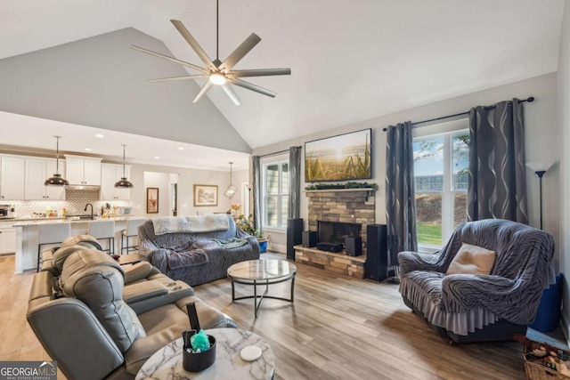 living room with a stone fireplace, ceiling fan, sink, high vaulted ceiling, and light hardwood / wood-style flooring