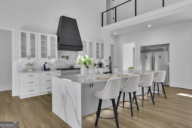 kitchen featuring white cabinetry, custom exhaust hood, stainless steel appliances, a kitchen island with sink, and a kitchen breakfast bar