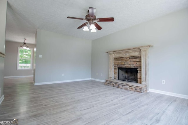 unfurnished living room with light wood-type flooring, ceiling fan with notable chandelier, a stone fireplace, and a textured ceiling