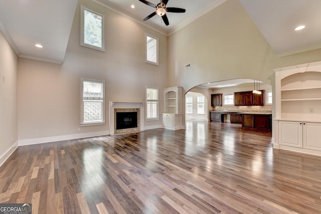 unfurnished living room featuring a towering ceiling, hardwood / wood-style flooring, ornamental molding, built in features, and ceiling fan