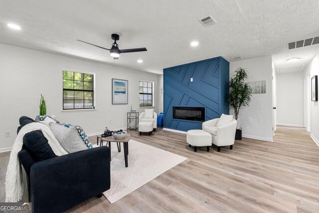 living room with ceiling fan, a textured ceiling, a large fireplace, and light wood-type flooring