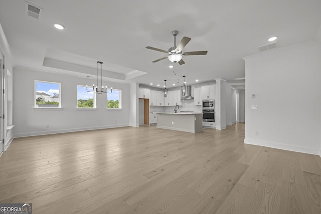 unfurnished living room with ceiling fan, a chandelier, recessed lighting, light wood-style flooring, and a tray ceiling