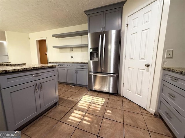 kitchen with a textured ceiling, stainless steel refrigerator with ice dispenser, light tile patterned floors, gray cabinetry, and stone counters