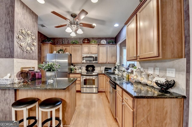 kitchen with appliances with stainless steel finishes, dark stone countertops, light hardwood / wood-style floors, a kitchen breakfast bar, and ceiling fan