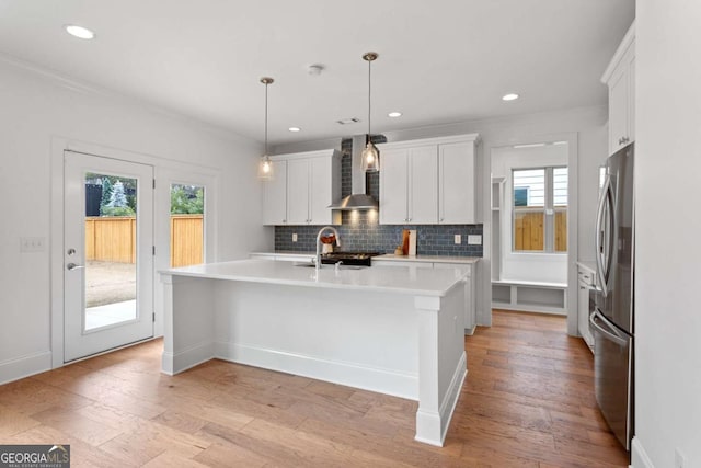 kitchen featuring white cabinets, wall chimney range hood, hanging light fixtures, a kitchen island with sink, and stainless steel refrigerator