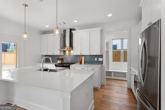 kitchen with white cabinets, wall chimney exhaust hood, decorative light fixtures, stainless steel appliances, and an island with sink