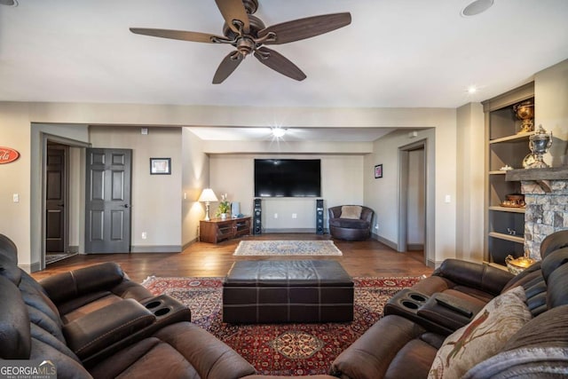 living room with hardwood / wood-style flooring, ceiling fan, built in features, and a stone fireplace