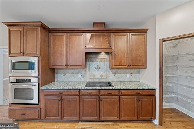 kitchen featuring custom exhaust hood, appliances with stainless steel finishes, decorative backsplash, light hardwood / wood-style flooring, and light stone counters