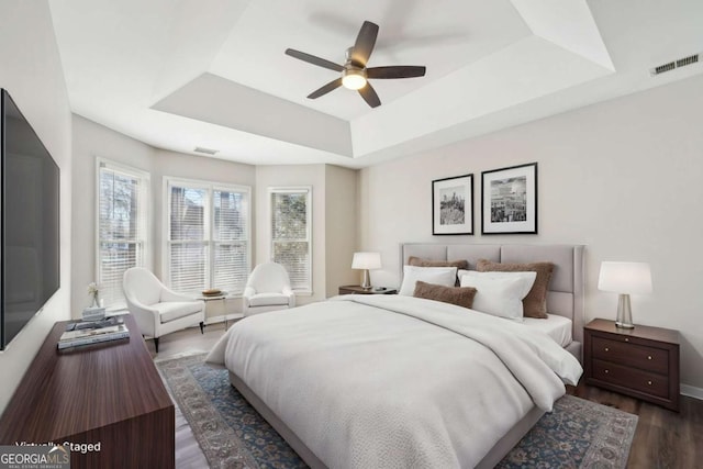 bedroom with ceiling fan, a raised ceiling, dark hardwood / wood-style floors, and multiple windows