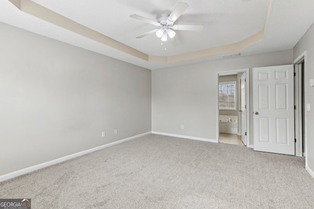 unfurnished bedroom with a tray ceiling, ensuite bath, light colored carpet, and ceiling fan