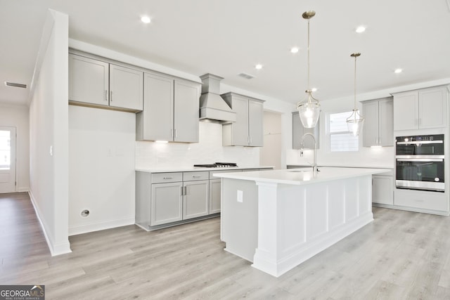 kitchen with custom exhaust hood, stainless steel double oven, sink, an island with sink, and gray cabinets