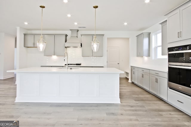 kitchen with pendant lighting, double oven, custom range hood, gray cabinetry, and a center island with sink