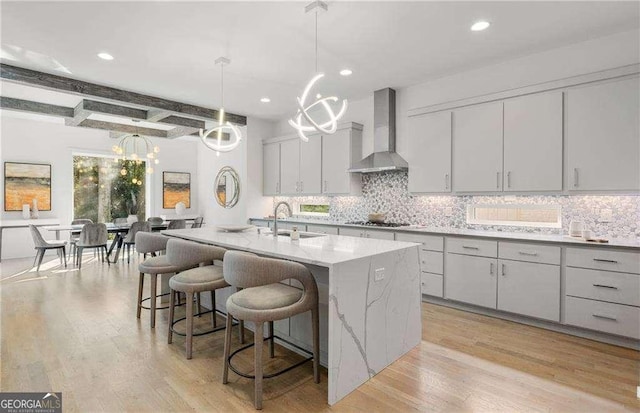 kitchen with decorative light fixtures, wall chimney range hood, sink, a kitchen island with sink, and beam ceiling