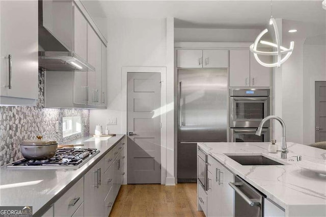 kitchen featuring appliances with stainless steel finishes, hanging light fixtures, sink, light stone counters, and wall chimney exhaust hood