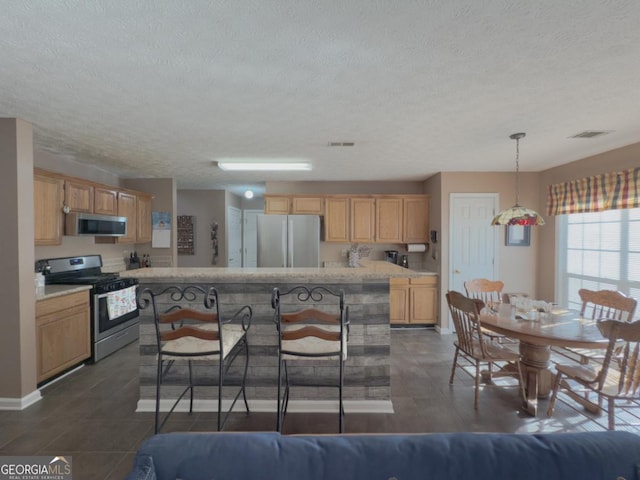 kitchen featuring a textured ceiling, appliances with stainless steel finishes, a center island, decorative light fixtures, and light brown cabinets