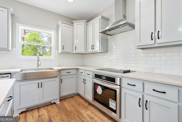 kitchen with oven, black electric stovetop, white cabinetry, sink, and wall chimney exhaust hood