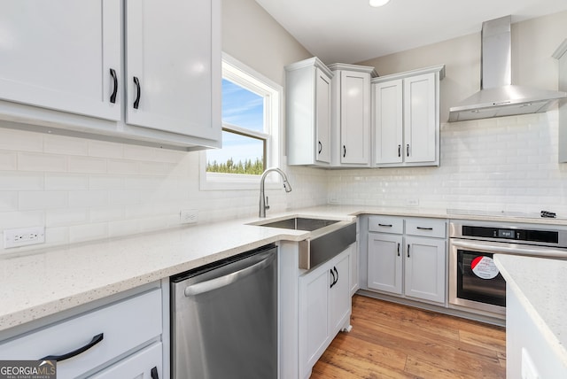 kitchen with appliances with stainless steel finishes, wall chimney range hood, white cabinets, sink, and light hardwood / wood-style flooring