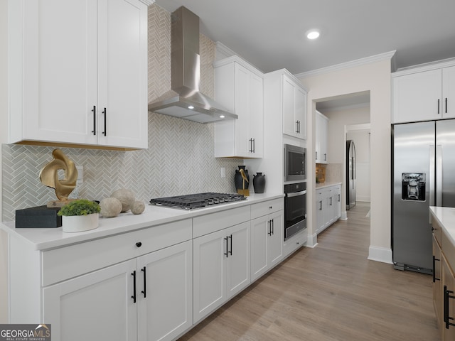 kitchen featuring wall chimney range hood, ornamental molding, stainless steel appliances, decorative backsplash, and white cabinets