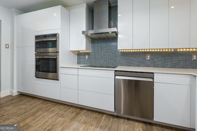 kitchen featuring white cabinets, decorative backsplash, stainless steel appliances, wall chimney range hood, and light hardwood / wood-style flooring