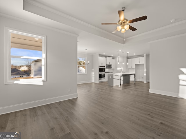 unfurnished living room with dark wood-type flooring, sink, crown molding, a tray ceiling, and ceiling fan with notable chandelier