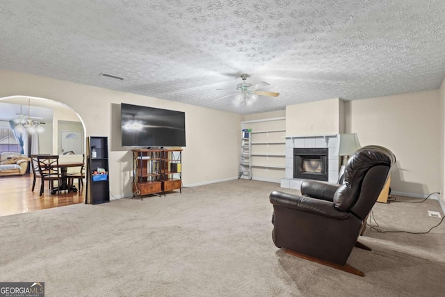 carpeted living room with a tiled fireplace, ceiling fan with notable chandelier, and a textured ceiling