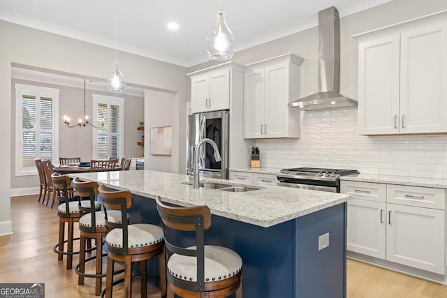 kitchen with hanging light fixtures, appliances with stainless steel finishes, wall chimney exhaust hood, sink, and a kitchen island with sink