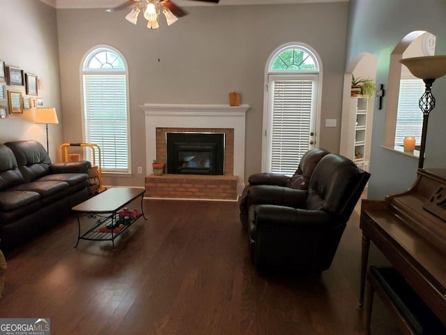 living room with a brick fireplace, dark wood-type flooring, and ceiling fan