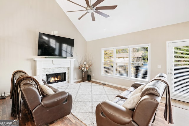 living room with ceiling fan, wood-type flooring, a stone fireplace, and high vaulted ceiling