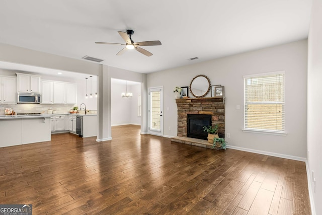 unfurnished living room with dark wood-type flooring, sink, a stone fireplace, and ceiling fan with notable chandelier