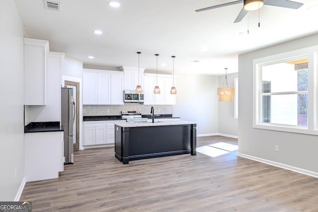 kitchen with appliances with stainless steel finishes, white cabinets, backsplash, hanging light fixtures, and a kitchen island with sink